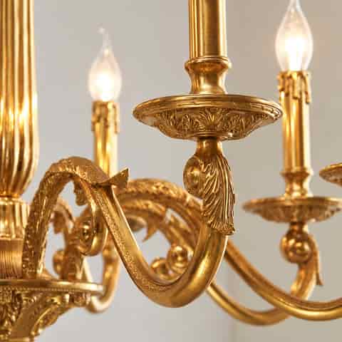 Close up of an ornate gold chandelier with carved leaf scrollwork, fluted details and decorative candle style light arms